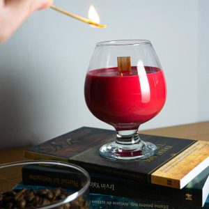 lumânare parfumată din ceară naturala de soia cu fitil din lemn în pahar de sticlă, așezat pe niște cărți și cu o mână ce ține un chibrit aprins